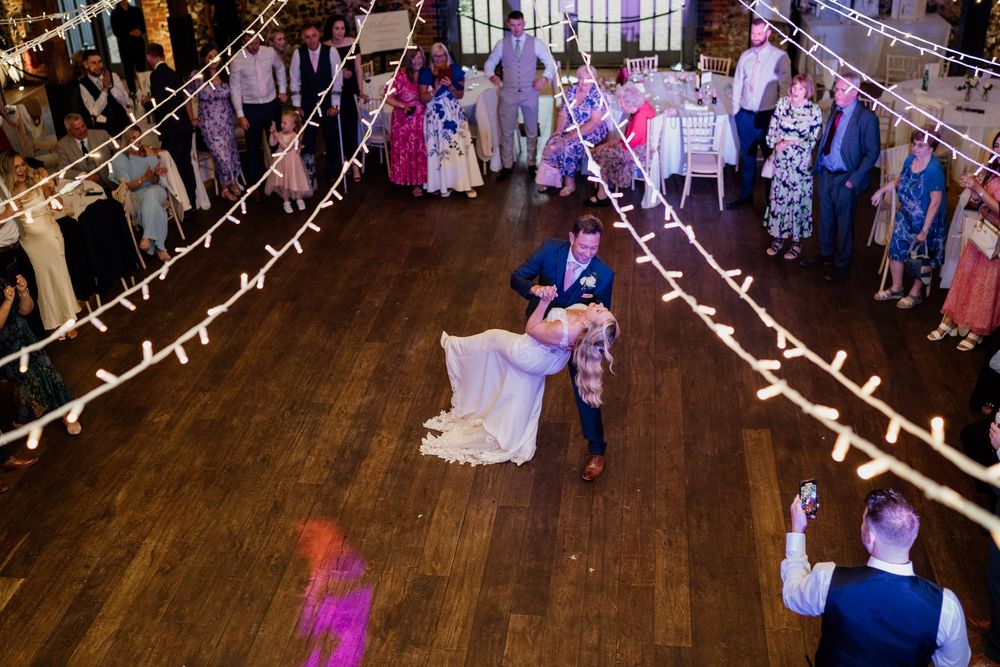 A couple shares their first dance at Oxnead Hall on a wooden floor beneath string lights while wedding guests watch from the perimeter.