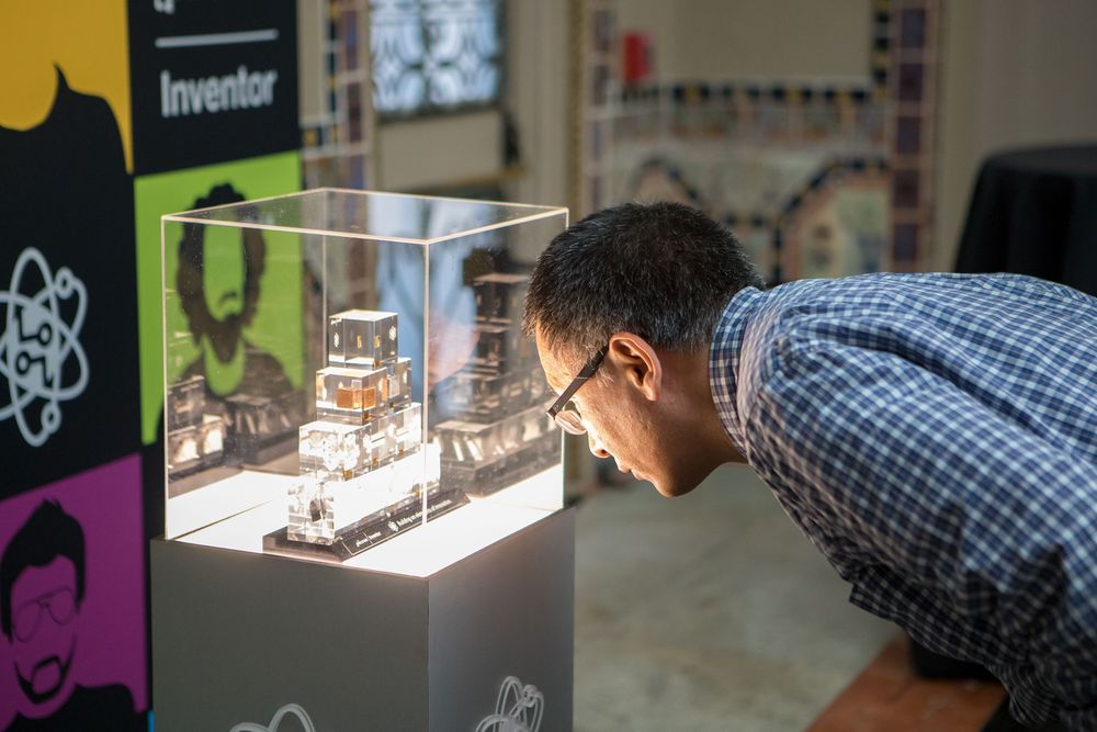 Someone leans in to examine a detailed miniature exhibit displayed in a clear museum case with colorful wall art behind.