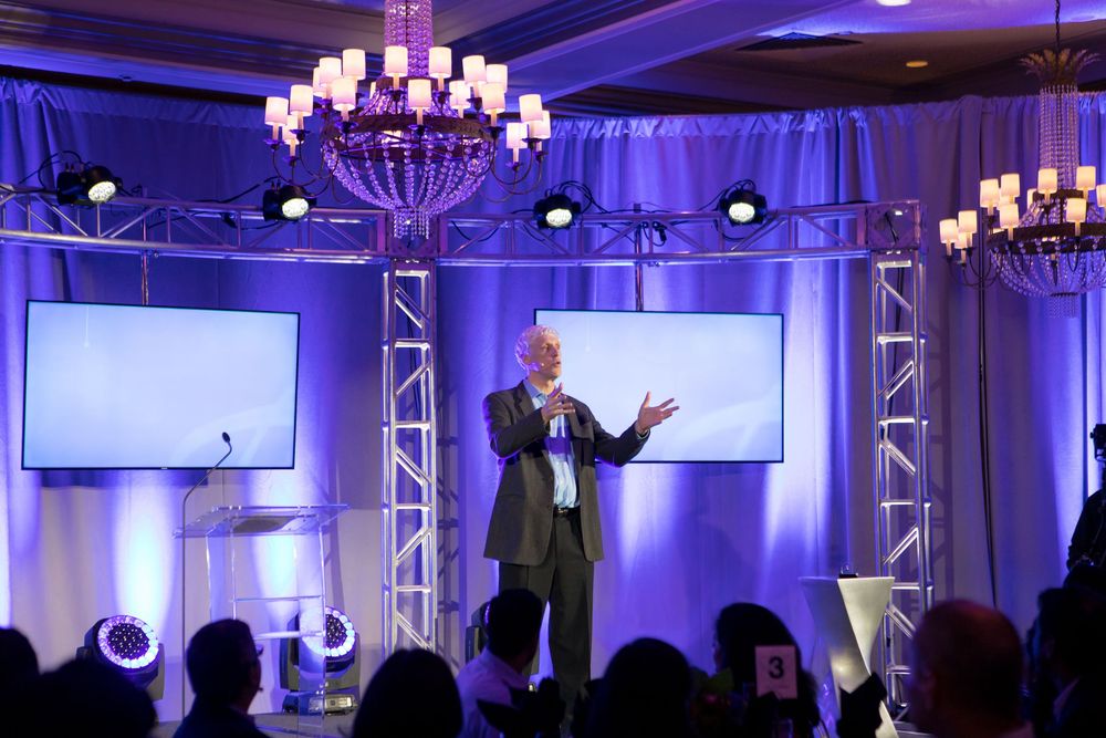 Speaker presenting to audience in elegant ballroom with blue lighting and crystal chandeliers during corporate event.