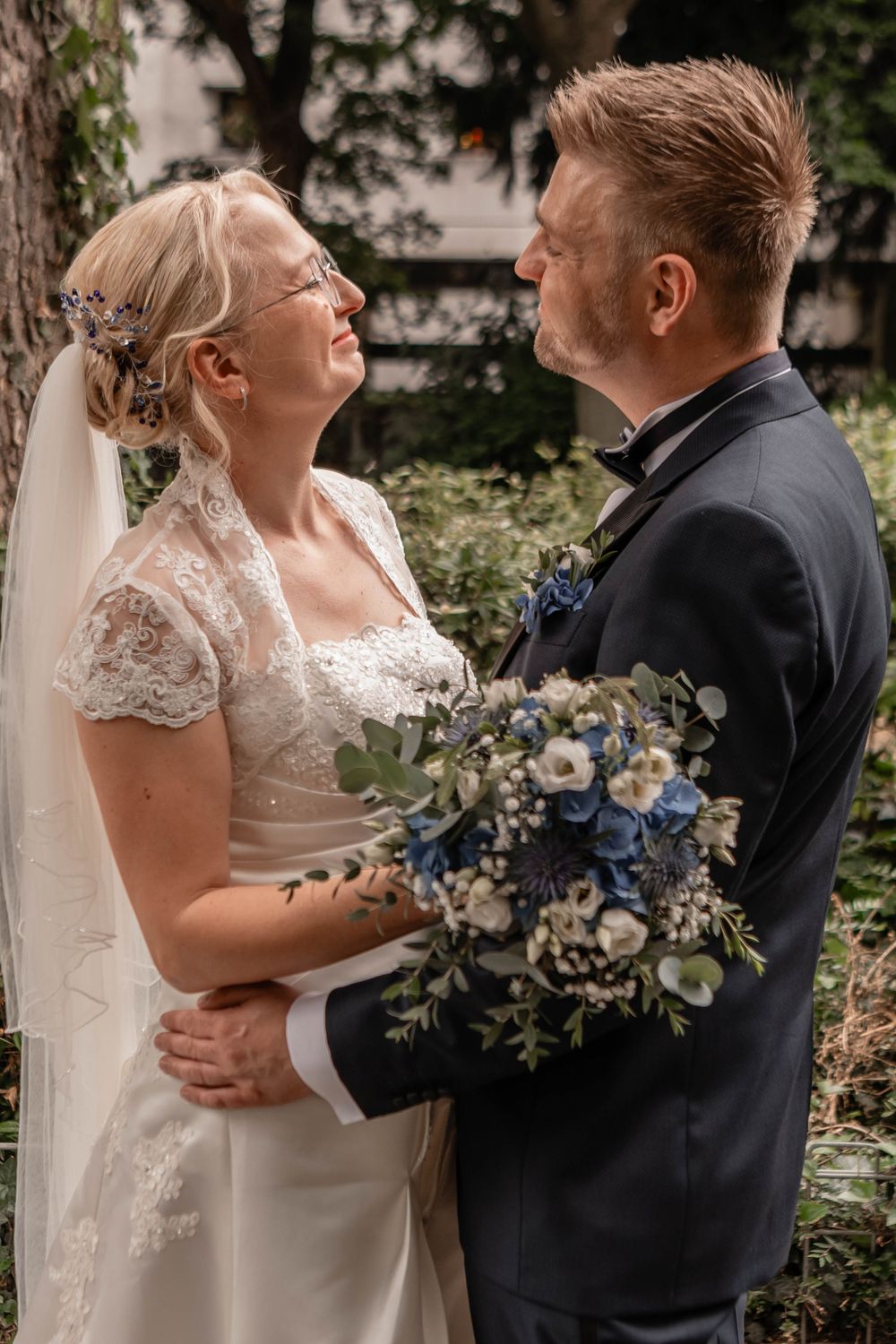 A bride and groom share an intimate moment outdoors, with the bride holding a blue and white floral bouquet.