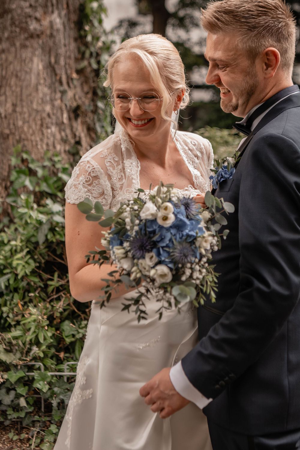 Candid wedding moment captures couple laughing together while holding a blue and white floral bouquet in garden setting.