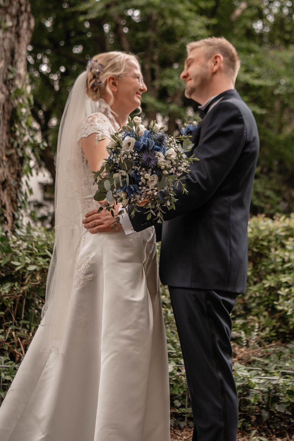 A bride and groom share a joyful moment outdoors with a stunning blue and white floral bouquet at their wedding.