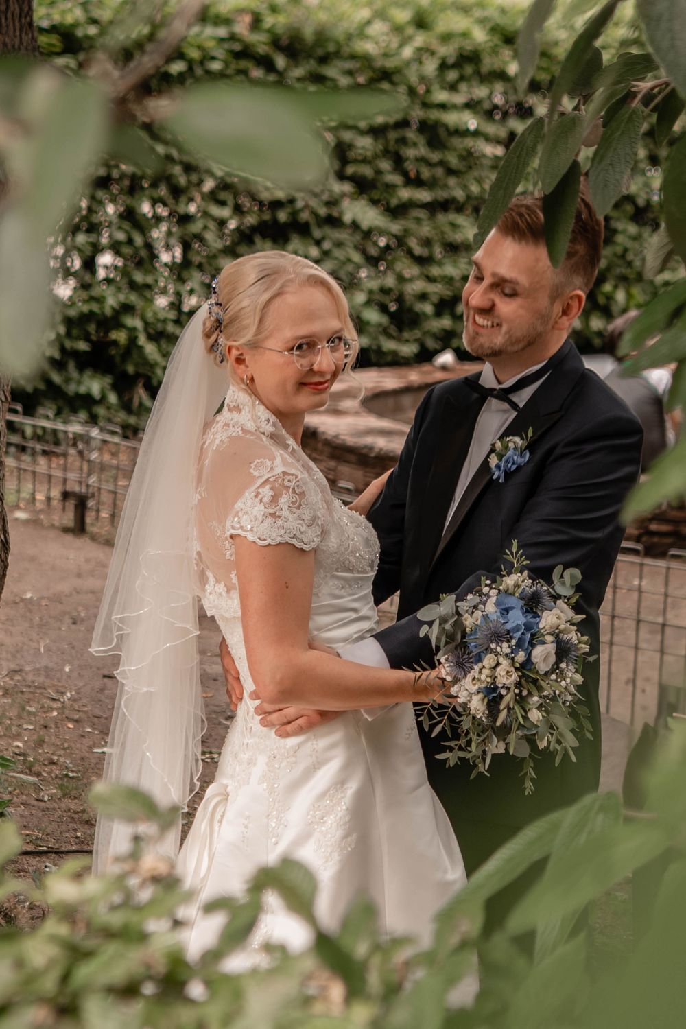 Newlywed couple sharing a romantic moment in an outdoor garden setting with blue floral bouquet and lace details.