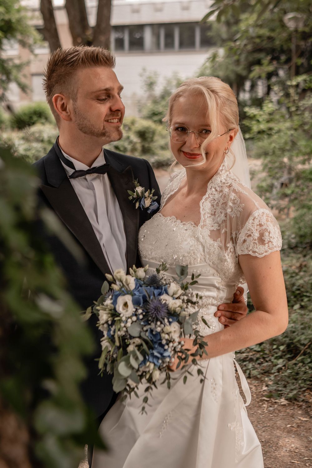 A bride and groom share a romantic moment outdoors with a stunning blue and white wedding bouquet in a garden setting.