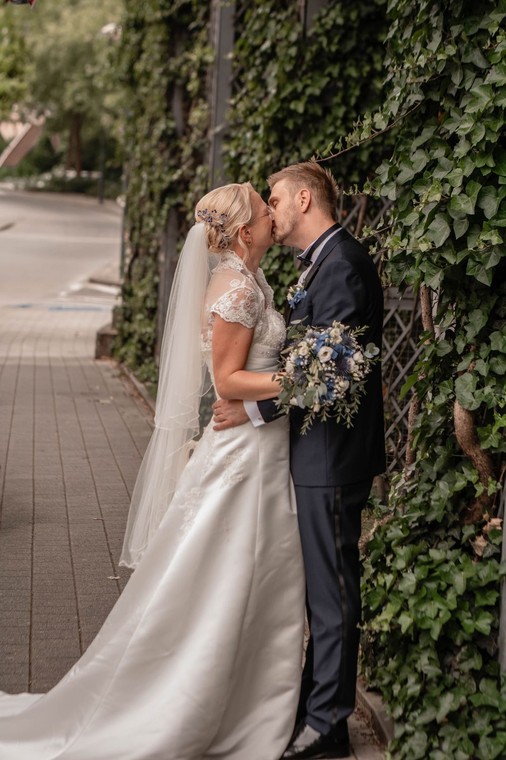 A romantic wedding scene with a bride in a flowing white gown and groom in dark suit embracing against an ivy-covered wall.