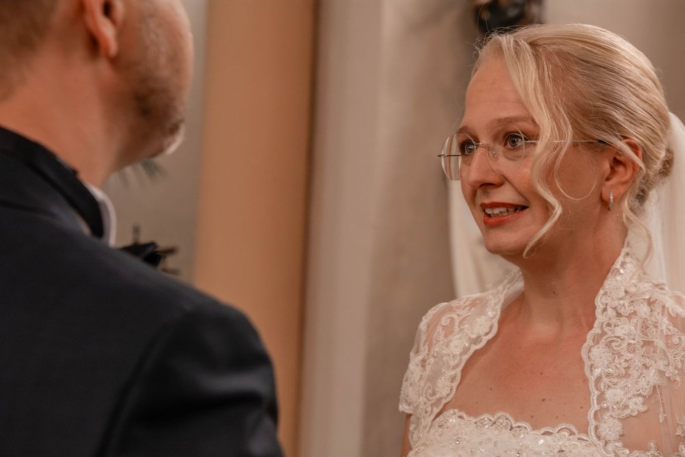 A bride in a white lace wedding dress and glasses smiles while looking at her partner during their wedding ceremony.