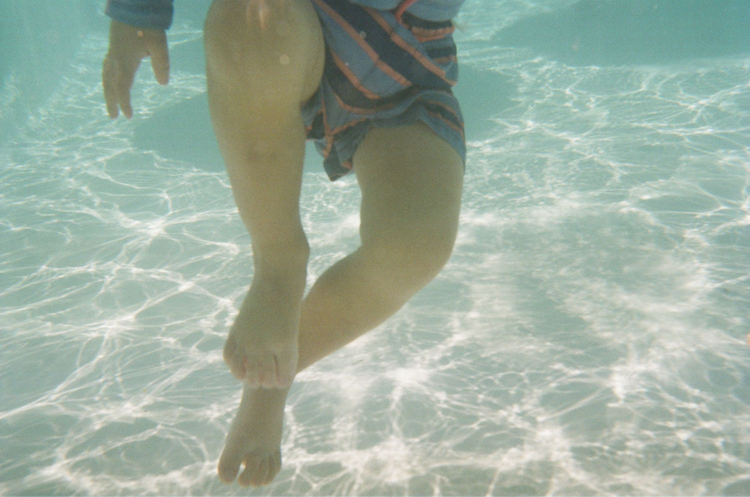 Underwater view of legs in striped swim shorts floating in clear turquoise pool water.