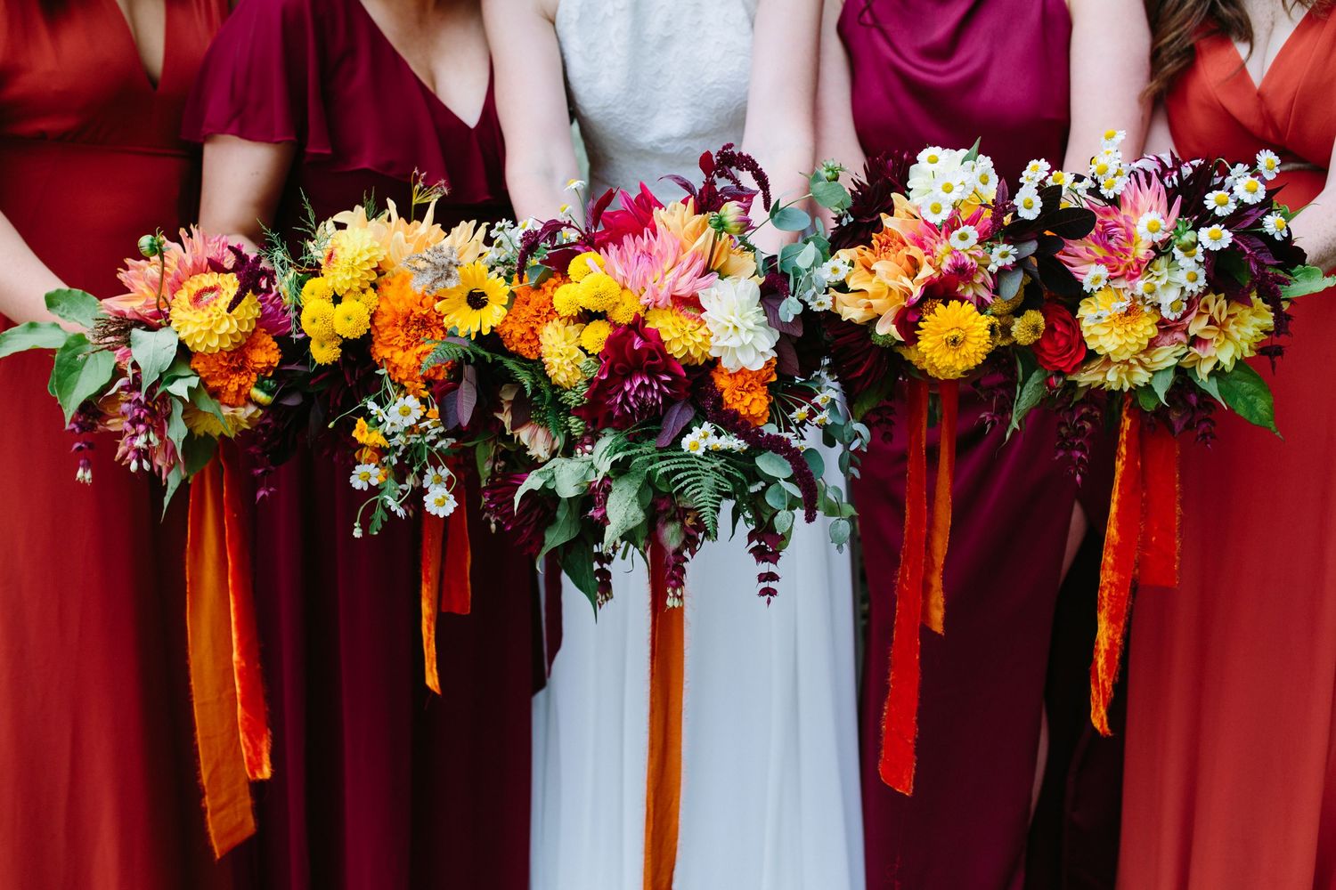 Wedding party holds vibrant autumn bouquets with sunflowers, pink blooms and orange ribbons against burgundy dresses.