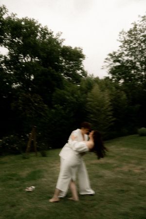 A couple in white attire shares a romantic dip kiss on a lush green lawn surrounded by trees.