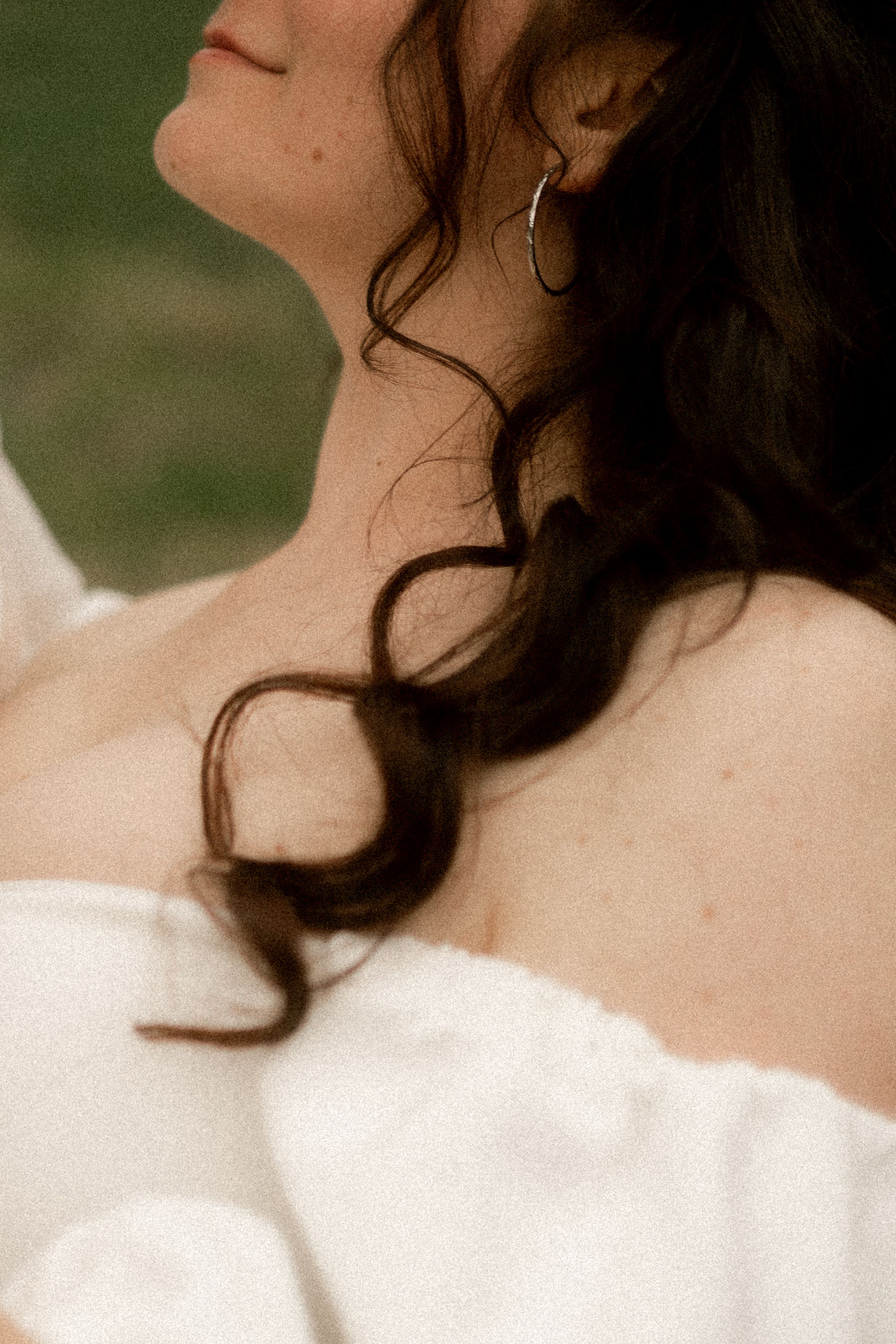 Close-up of bride with dark curly hair, hoop earring, and off-shoulder white dress gazing upward softly.