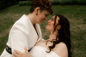 Two brides share a tender moment, smiling at each other in a lush green outdoor setting.