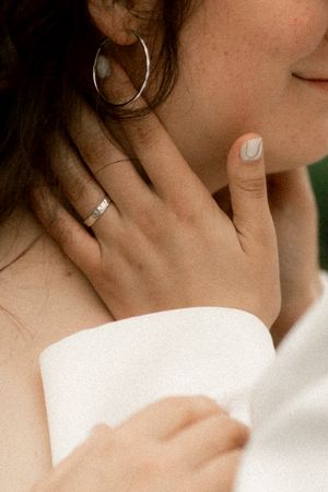 Close-up of bride wearing silver hoop earring and wedding band ring, with manicured nails against white off-shoulder dress.