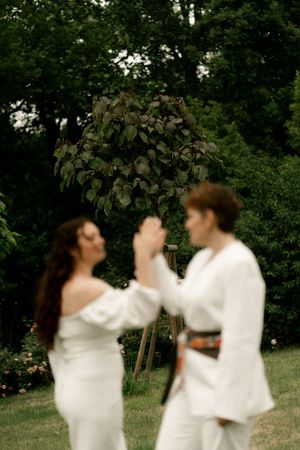 Two brides in white dresses dancing under leafy arch during outdoor garden wedding ceremony.