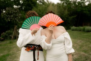 Two brides in white holding colorful green and orange fans covering their faces outdoors.