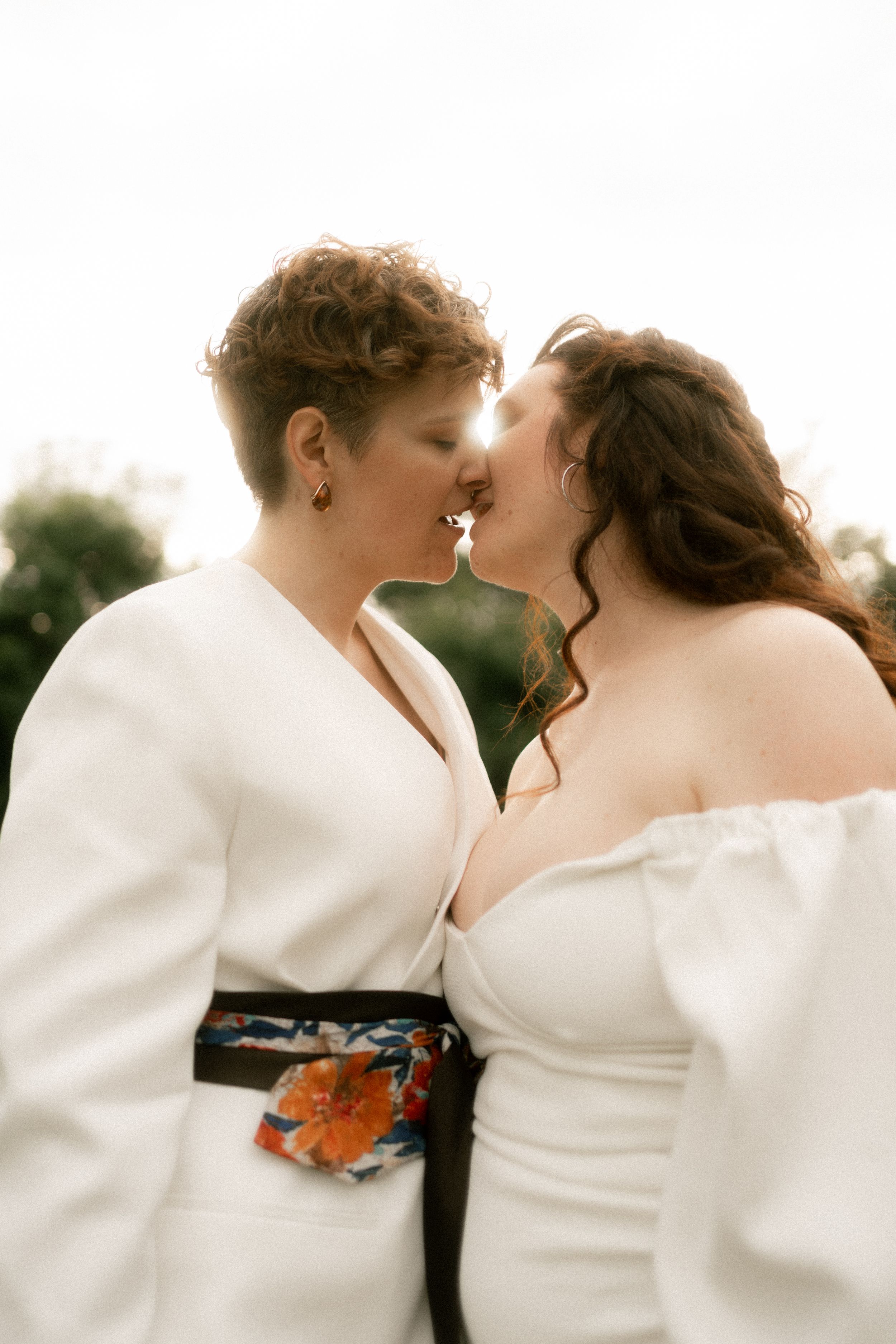 Two brides in white outfits sharing a tender kiss outdoors on their wedding day.