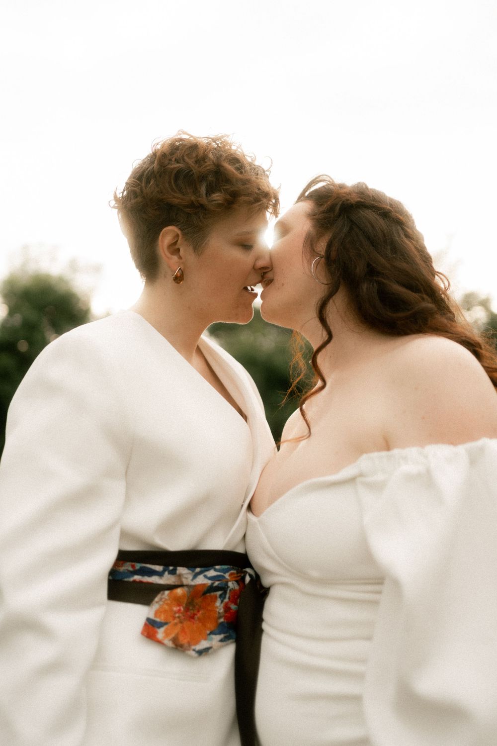 Two brides in white outfits sharing a tender kiss outdoors on their wedding day.