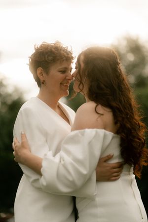 Two brides in white embrace and smile at golden hour in a romantic outdoor wedding portrait.