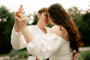 Two brides in white dancing closely together outdoors, sharing an intimate moment in a lush green setting.
