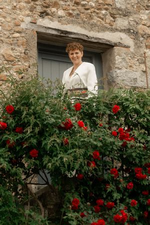 Woman in white top smiling among red roses by a rustic stone building with a blue window.