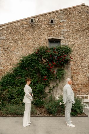 Two people in matching light suits stand apart before a rustic stone wall adorned with climbing red roses.