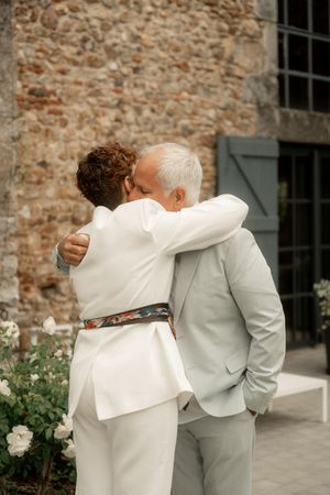 Two people in elegant suits share a warm embrace outdoors near a stone building with white roses.