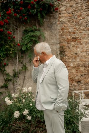 Emotional elderly man in light gray suit wiping tears near stone wall with red roses at wedding.