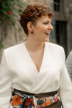 Woman with short curly hair laughing in white wrap top with floral sash at outdoor event.