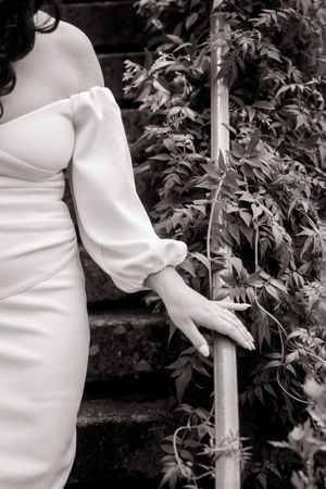 Black and white photo of a bride in an off-shoulder white gown holding a railing on stone steps beside lush greenery.