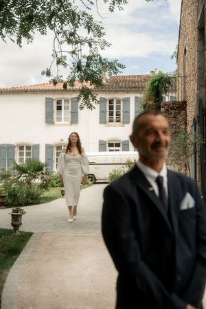 Bride in white dress walking toward groom in suit outside French countryside stone villa.
