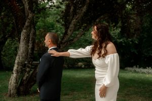 Bride in white off-shoulder gown touches groom's shoulder during outdoor first look among lush green trees.