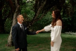 Bride and groom holding hands outdoors in lush garden, groom in black suit, bride in off-shoulder white gown.