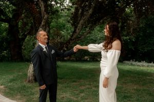 Smiling groom in black suit holds hands with bride in elegant white off-shoulder dress in a green garden.