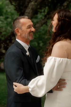 Smiling groom in black suit embracing bride in off-shoulder white wedding dress outdoors.