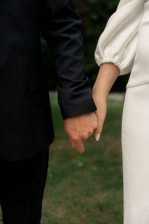 Close-up of a bride and groom holding hands, wearing white and black wedding attire outdoors.