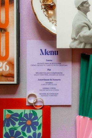 Elegant wedding menu card in lavender with French text, surrounded by rings, florals, and colorful stationery.