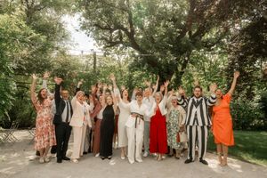 Joyful wedding guests celebrating outdoors with raised hands under large trees in a festive garden setting.