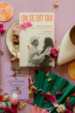 French wedding invitation reading On Se Dit Oui surrounded by dried flowers, rose petals, rings, and a menu card on pink background.