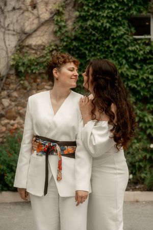 Two brides in matching white outfits sharing a tender moment against an ivy-covered stone wall.