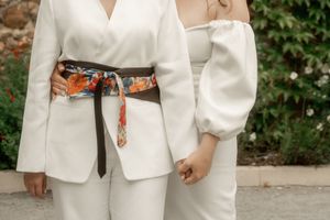 Two brides in white outfits hold hands, one wearing a floral obi belt over a white blazer suit.