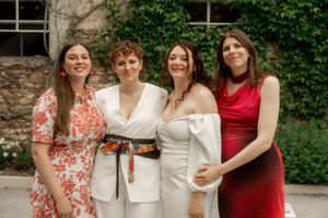 Four women smiling together outdoors, one in white wedding dress with colorful sash.