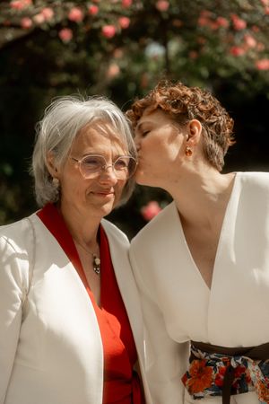 A young woman with curly hair kisses an older gray-haired woman wearing glasses outdoors near blooming flowers.