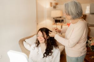 Older woman with gray hair styling the dark curly hair of a smiling young bride in a white robe.