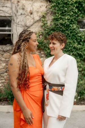 Two women laughing joyfully outdoors, one in orange dress and one in white suit at wedding.