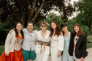 Six women smile together outdoors at a garden celebration, dressed in colorful and elegant attire.
