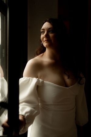 Bride in white off-shoulder gown gazes out window in dramatic moody lighting.