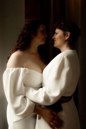 Two brides in white dresses embrace and smile at each other in a romantic wedding portrait.