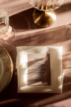 Elegant wedding menu card on white linen napkin with gold candlestick and floral china plate in warm sunlight.