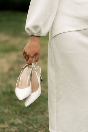 Bride in white dress holding elegant white pointed-toe slingback heels outdoors on green grass.