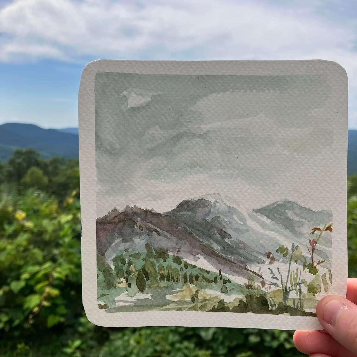 A hand-painted watercolor of a misty mountain scene with rolling hills and soft greenery held against a real life mountain backdrop