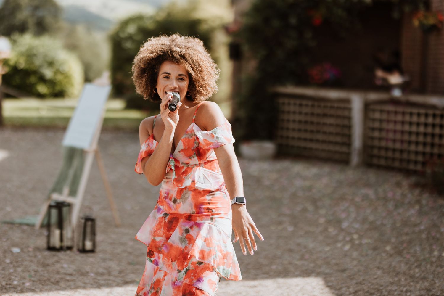A woman in a flowing coral floral dress walks through a pebbled garden area.